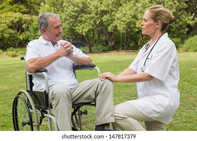 Cheerful Man In A Wheelchair Talking With His Nurse Kneeling Beside Him In The Park On A Sunny Day