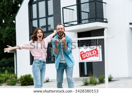 cheerful man and woman celebrating near house and board with sold letters 