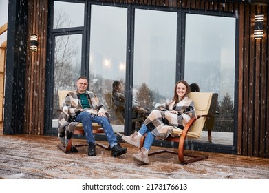 Cheerful Man And Woman Wrapped In Blanket Sitting In Chairs Outside Scandinavian House Barnhouse. Happy Couple Resting Together Outdoors Near Building With Panoramic Window Under Winter Snow.