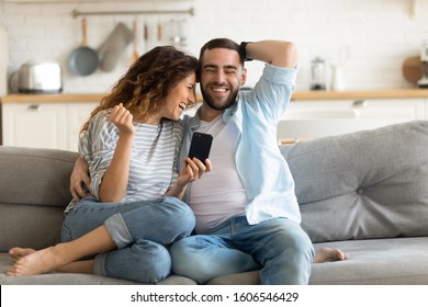 Cheerful Married Couple Resting On Couch Enjoy Lazy Weekend Together At Home Having Fun Using Online Amusing Apps, Wife Holding Smart Phone Spouses Laughing On Funny Video, Website With Pranks Concept