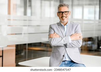 Cheerful mature 50s businessman, startup owner, investor standing with arms crossed in his office. Mature proud gray-haired male boss, ceo, professional in formal wear looking at camera and smiling