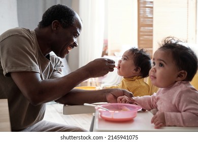 Cheerful mature African American father taking care of his baby daughters feeding them with fruit puree - Shutterstock ID 2145941075