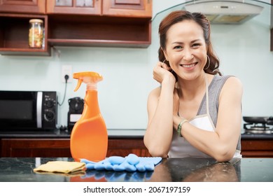 Cheerful mature Asian woman resting at kitchen table with bottle of cleaning detergent and rubber gloves - Powered by Shutterstock