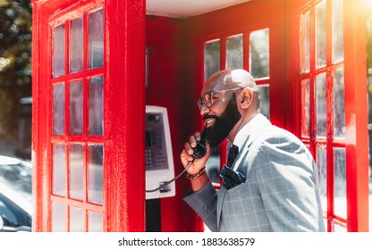 A Cheerful Mature Bald Elegant Black Man Entrepreneur In A Fashionable Business Costume, Eyeglasses, And With A Well-groomed Beard Is Speaking On A Telephone Standing In A Retro Red Phone-booth