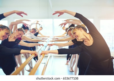 Cheerful mature ballerinas stretching with hands up standing at mirror in ballet class.