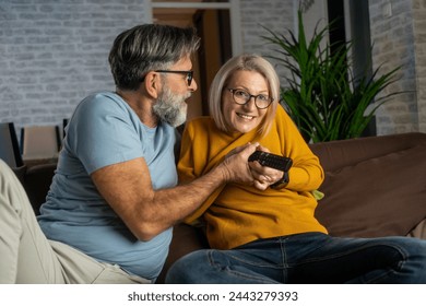 Cheerful mature beautiful love couple fighting for tv remote while sitting on the sofa in living room. Happy couple sitting on couch at home watching tv - Powered by Shutterstock