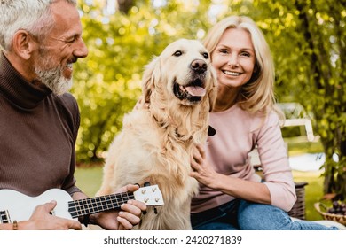 Cheerful mature caucasian couple spending time together with their dog golden retriever while grey-haired pet owner husband man playing the ukulele in park - Powered by Shutterstock