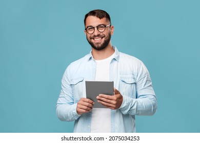 Cheerful mature caucasian guy in glasses with tablet look at camera, isolated on blue background, free space. Modern businessman, blogger, teacher with device, new normal, freelance and work remotely