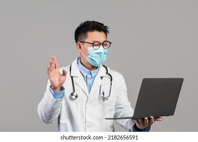 Cheerful Mature Chinese Man Doctor In White Coat, Glasses, Protective Mask With Stethoscope Waving Hand, Looking At Laptop, Isolated On Gray Background, Studio. Video Call, Online Consultation Remote