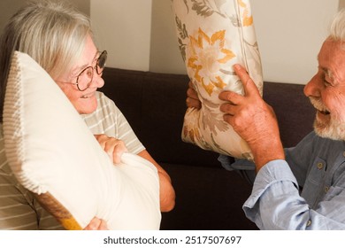 Cheerful mature couple enjoying a lively pillow fight on the couch at home. Despite their senior years, they exhibit youthful energy and playfulness, showcasing a loving and fun-filled life together. - Powered by Shutterstock