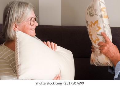 Cheerful mature couple enjoying a lively pillow fight on the couch at home. Despite their senior years, they exhibit youthful energy and playfulness, showcasing a loving and fun-filled life together. - Powered by Shutterstock