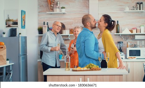 Cheerful Mature Couple Hugging Flirting In Kitchen Infront Senior Parents, Drinking A White Wine. Young Family Romantic, Funny, Having Tenderness And Affection Moment In Modern, Luxury Dining Room