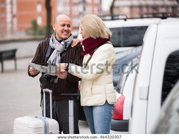 Cheerful  mature couple with luggage standing\
at street and checking\
direction\
