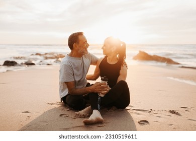 Cheerful mature european husband hugs wife in sportswear, enjoy workout with bottle of water together in morning, sit on sea beach, outdoor. Rest from sports, fitness and body care - Shutterstock ID 2310519611