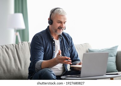 Cheerful mature grey-haired man freelancer working from home during COVID-19 pandemic, having video chat with employer or clients, using laptop, headset and notepad, copy space