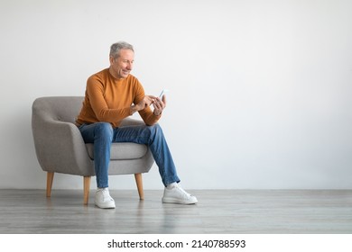 Cheerful Mature Guy Using Mobile Phone Testing New Application Sitting In Armchair Isolated On White Studio Wall Background. Gadgets And Mobile Communication Concept. Free Empty Copy Space For Text - Powered by Shutterstock