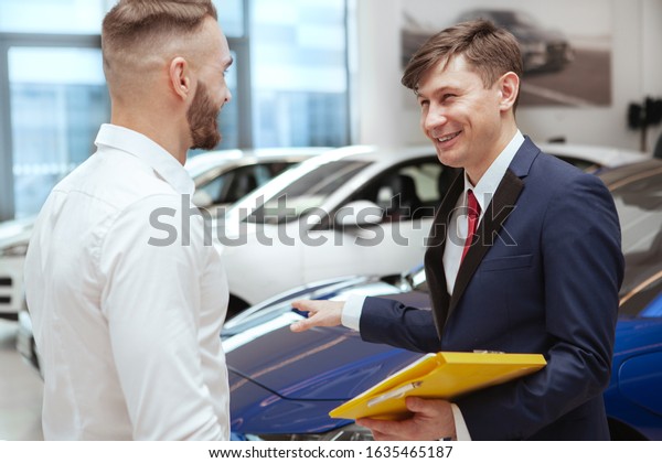 Cheerful mature salesman laughing, talking to\
a client at car\
dealership