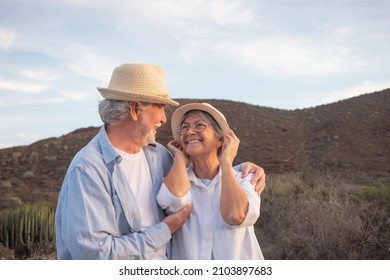 Cheerful mature senior couple enjoying healthy activity in outdoor mountain excursion at sunset light looking each other in the eyes hugging. Smiling handsome retired wearing hats. Copy space - Powered by Shutterstock