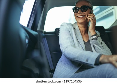 Cheerful Mature Woman In Business Suit Sitting On Backseat Of Her Car And Talking On Mobile Phone. Senior Businesswoman Using Phone While Traveling By A Car.