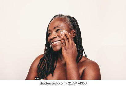 Cheerful mature woman with dreadlocks applying moisturizing cream on her face. Smiling dark-skinned woman taking care of her flawless melanated skin. Happy black woman ageing gracefully.