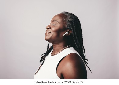 Cheerful mature woman with dreadlocks listening to some music on bluetooth earbuds. Happy middle-aged woman enjoying her favourite playlist while standing against a studio background. - Powered by Shutterstock