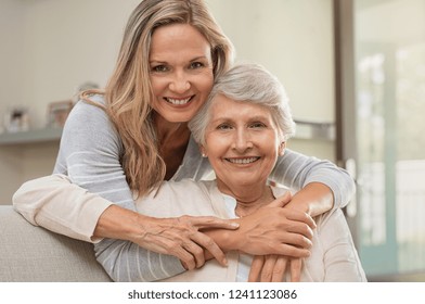 Cheerful Mature Woman Embracing Senior Mother At Home And Looking At Camera. Portrait Of Elderly Mother And Middle Aged Daughter Smiling Together. Happy Daughter Embracing From Behind Elderly Mom.