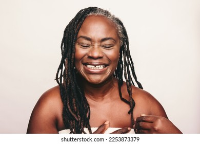 Cheerful mature woman laughing happily while wrapped in a bath towel. Refreshed woman with dreadlocks standing against a white background. Joyful middle aged woman taking care of her ageing body.