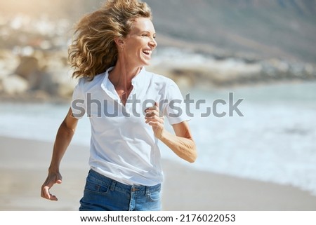 Cheerful mature woman running on the beach on a sunny day. Beautiful middle aged woman laughing, being active and having fun during summer vacation. Energetic lady spending her free time by the sea