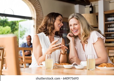 Cheerful mature women enjoying a funny video on mobile phone. Mature friends reading a funny message over smartphone. Mid woman showing a cellphone to her friend while laughing over breakfast.