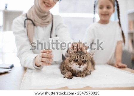 Cheerful medical person in hijab putting pill container near mature cat on desk in vet office with caucasian girl alongside. Arabian animal doctor preventing flea infestation for domestic friend.