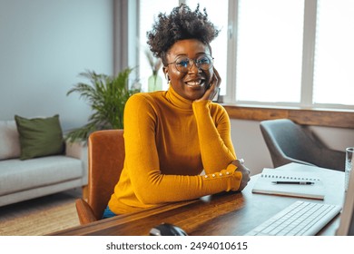 A cheerful mid-adult black businesswoman wearing glasses and a yellow turtleneck sits at her wooden desk equipped with a modern computer, notepad, and pen in a well-lit home office. - Powered by Shutterstock