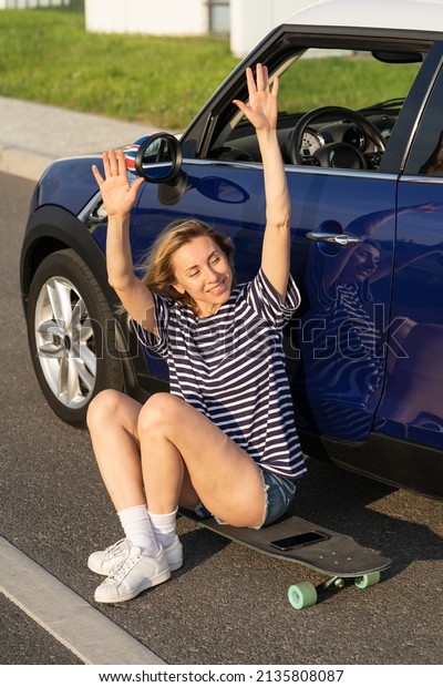 Cheerful middle age woman waving hand greeting\
welcoming friends while sitting on longboard outside modern car.\
Active adult female gathering with fellows to skateboarding in\
countryside on sunset