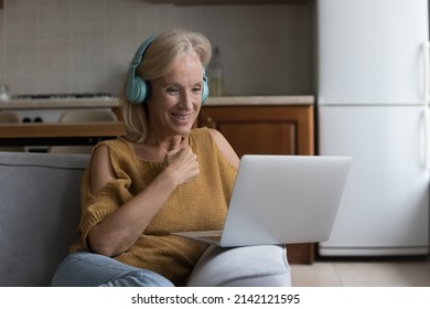 Cheerful middle aged 50s in big wireless headphones using laptop computer, talking to family, grandchildren on video call, listening learning audio, watching movie, sitting on sofa at home
