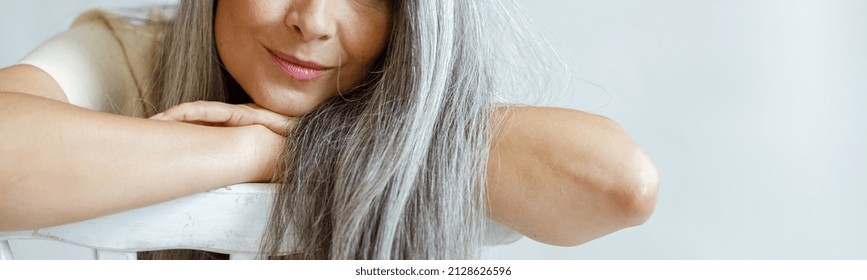 Cheerful middle aged Asian lady with flowing hoary hair sits backwards on chair on light background in studio. Mature beauty lifestyle
