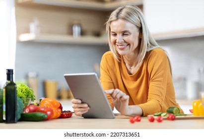 Cheerful middle aged blonde woman reading food blog on Internet and cooking healthy delicious meal, pretty lady standing next to kitchen table, using modern digital tablet, copy space - Powered by Shutterstock