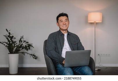 Cheerful Middle Aged Chinese Male Typing On Laptop, Have Online Chat Remotely, Sit In Armchair In Minimalist Room Interior. Video Call From Device, Work And Business And Social Distance Due Covid-19