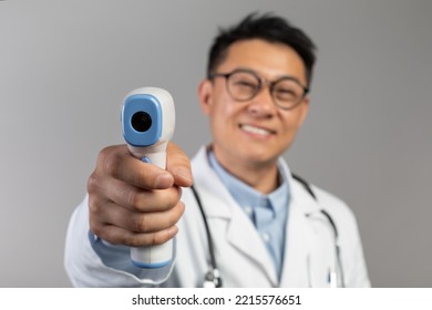 Cheerful Middle Aged Chinese Male Doctor In White Coat, Glasses Measures Temperature With Contactless Thermometer, Isolated On Gray Background, Studio. Diagnostics, Treatment Of Ill And Health Care