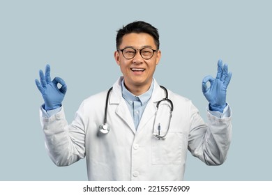 Cheerful Middle Aged Chinese Male Doctor In White Coat, Protective Gloves, Glasses Shows Ok Sign With Hands, Isolated On Blue Background. Great Treatment, Health Care Result, Medicine During Covid-19