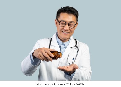 Cheerful Middle Aged Chinese Male Doctor In White Coat, Glasses Pour Out Pills From Jar On Hand, Isolated On Blue Background, Studio. Pharmacie, Therapist Recommendation, Ad And Offer, Health Care