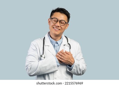 Cheerful Middle Aged Chinese Male Doctor In White Coat, Glasses Presses Hands To Chest Isolated On Blue Background, Studio. Gratitude, Love, Great Ad And Offer, Health Care And Medicine, Covid-19