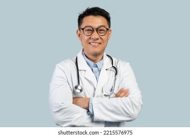 Cheerful Middle Aged Chinese Male Doctor In White Coat, Glasses With Crossed Arms Look At Camera, Isolated On Blue Background. Therapist Diagnostics, Ad And Offer, Treatment, Health Care And Medicine