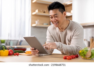 Cheerful Middle Aged Chinese Man Cooking Dinner At Home, Looking For Nice Recipe On Internet, Reading Food Blog, Using Modern Digital Tablet, Kitchen Interior, Copy Space. Cooking, Culinary Concept