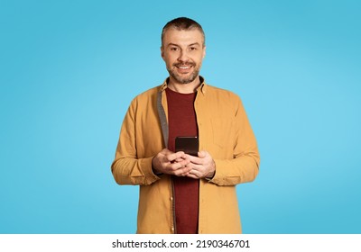 Cheerful Middle Aged Male Holding Smartphone Smiling To Camera Texting And Using Mobile Application Over Blue Background In Studio. Happy Man Posing With Cellphone. New App Advertisement