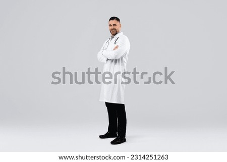 Cheerful middle aged man doctor posing with folded arms looking at camera standing on light background, wearing white uniform, coat and stethoscope, full body length, free space