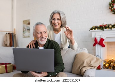 cheerful middle aged woman in glasses waving hand near husband during video call on christmas day - Powered by Shutterstock