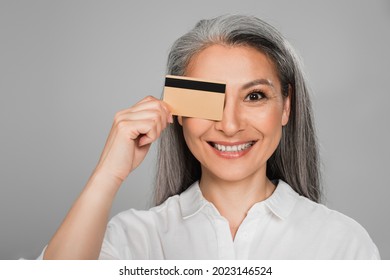 cheerful middle aged woman with grey hair obscuring eye with credit card isolated on grey - Shutterstock ID 2023146524