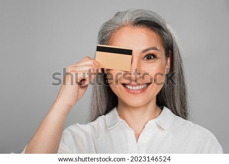 cheerful middle aged woman with grey hair obscuring eye with credit card isolated on grey