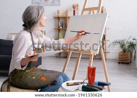 cheerful middle aged woman holding paintbrush and palette while painting on canvas