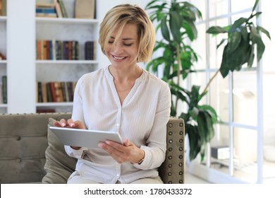 Cheerful Middle Aged Woman Sitting On Sofa, Using Computer Tablet Apps, Looking At Screen,reading Good News In Social Network, Shopping Or Chatting Online.