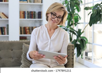 Cheerful Middle Aged Woman Sitting On Sofa, Using Computer Tablet Apps, Looking At Screen,reading Good News In Social Network, Shopping Or Chatting Online.